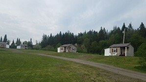 A rural setting with several small, single-story houses with attached porches, surrounded by a grassy field and dense forest. - 02) Northwoods Cabin (Stewartstown)
