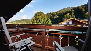 Eine Holzterrasse mit zwei Stühlen und einem Tisch, mit Blick auf einen bewaldeten Hang und ein Haus.