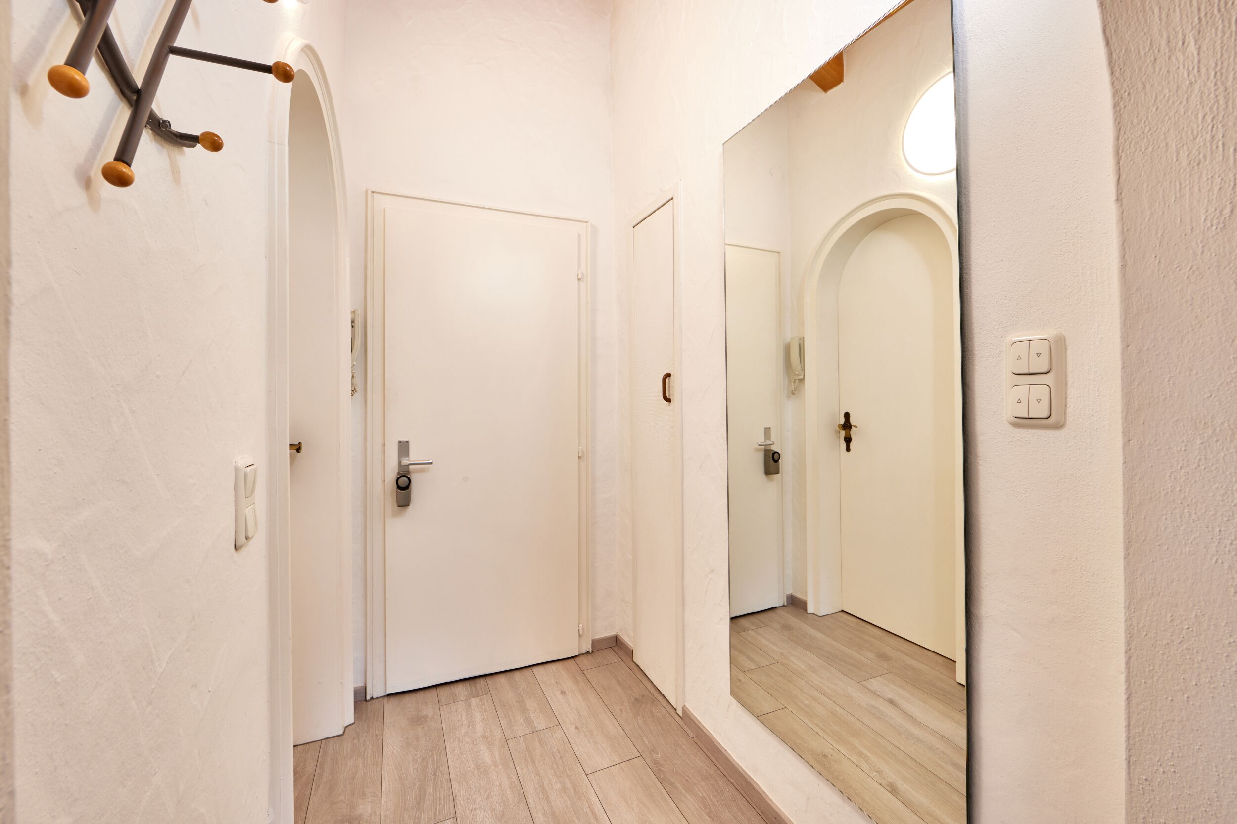 Un pasillo con paredes blancas, suelo de madera y un espejo que refleja una puerta y un interruptor de luz.