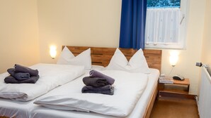 Un lit double avec du linge de lit blanc et deux oreillers, une tête de lit en bois, une table de chevet avec un téléphone et une lampe, et une fenêtre avec des rideaux bleus.