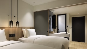 Una habitación de hotel moderna con dos camas grandes, un cabecero de madera, un espejo amplio, un escritorio empotrado con lavabo y una puerta oscura.