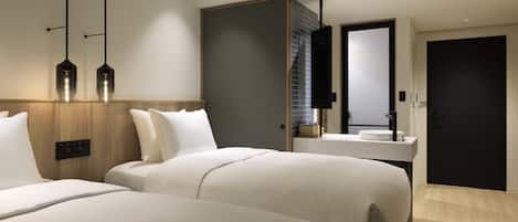 A modern hotel room with two large beds, a wooden headboard, a large mirror, a built-in desk with a sink, and a dark door.
