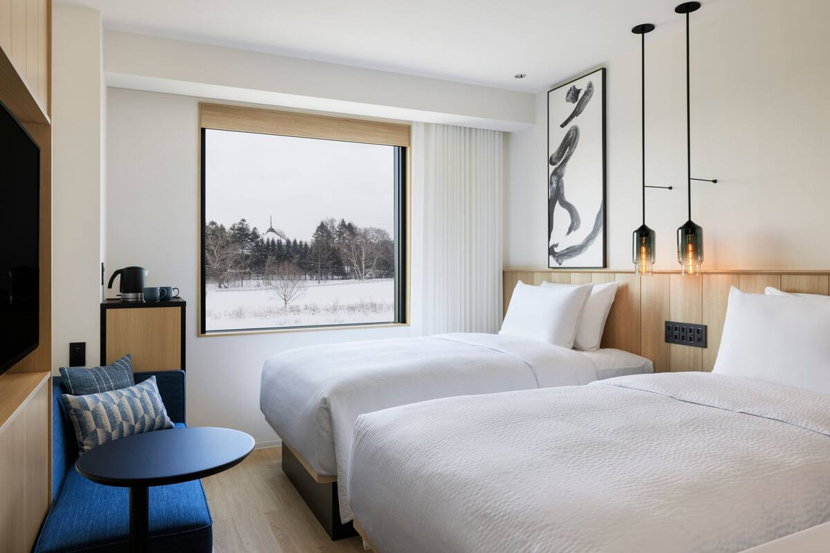 Habitación de hotel con dos camas, una mesita, un televisor y una ventana que ofrece vistas a árboles y un edificio.