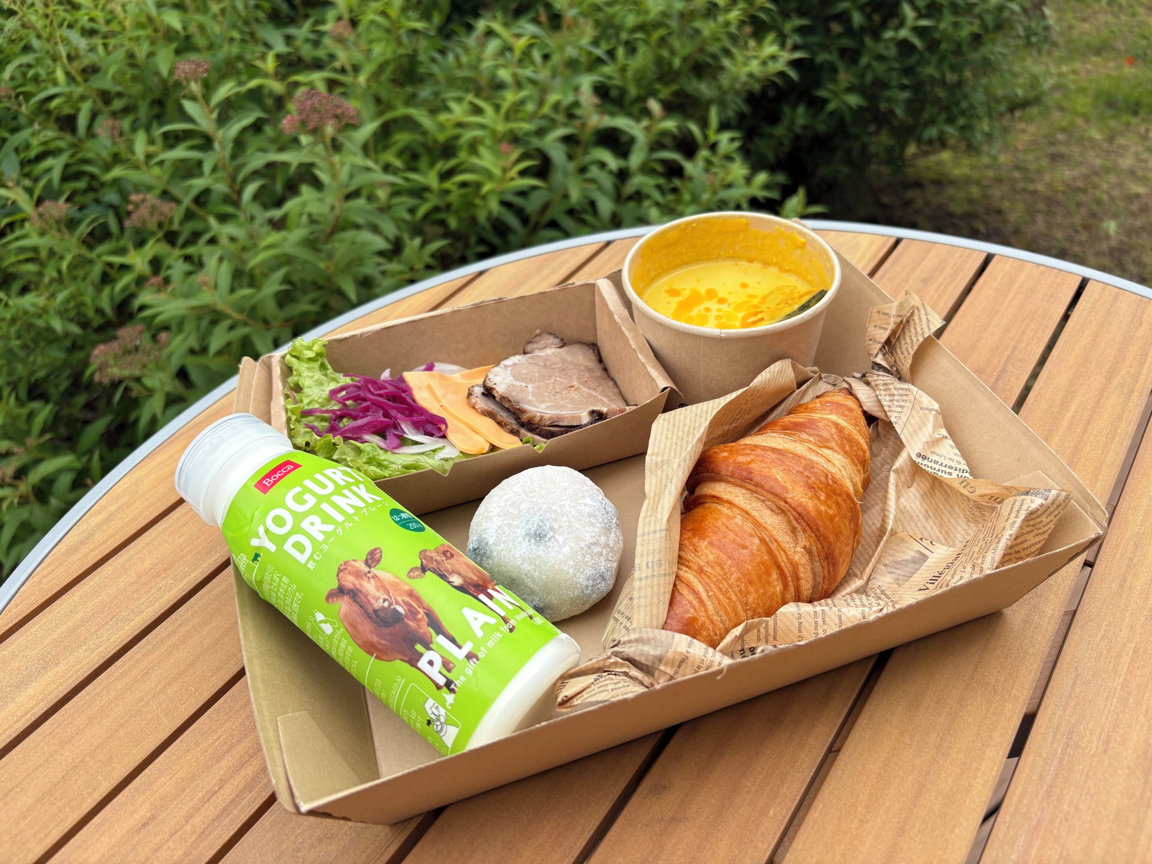 Comida con bebida de yogur, un croissant y una ensalada sobre una mesa de madera al aire libre.