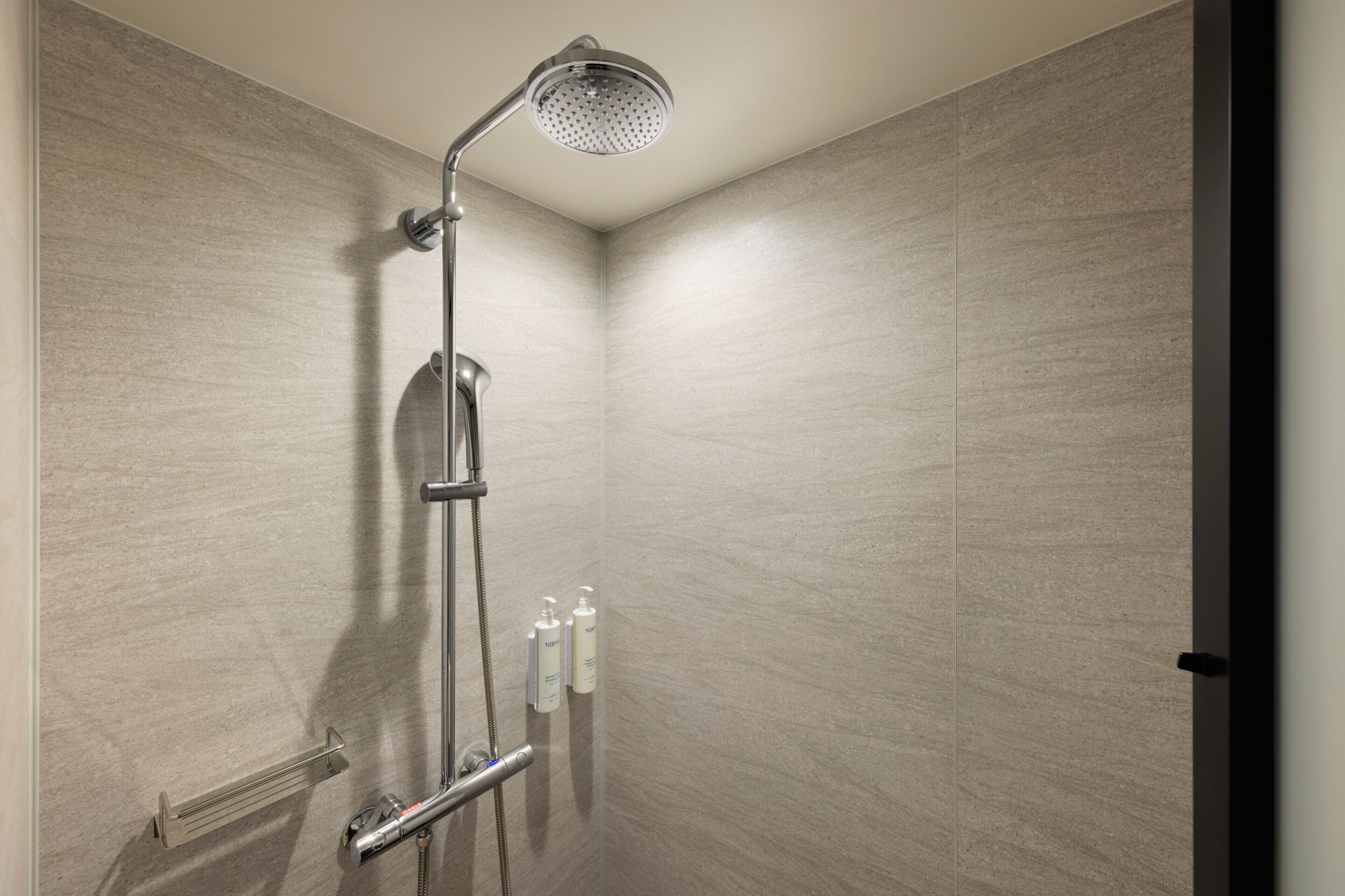 Une douche moderne équipée d’un pommeau de douche à main, d’un panneau de commande mural et de deux flacons de shampoing et d’après-shampoing.