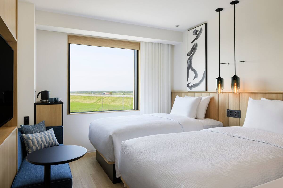 A hotel room with two beds, a small table, a sofa, a TV, and a large window offering a view of the countryside.