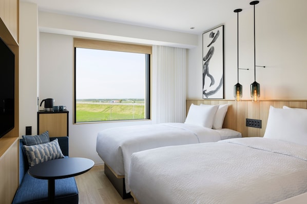 Une chambre d’hôtel avec deux lits, une petite table, un canapé, une télévision et une grande fenêtre donnant sur la campagne.
