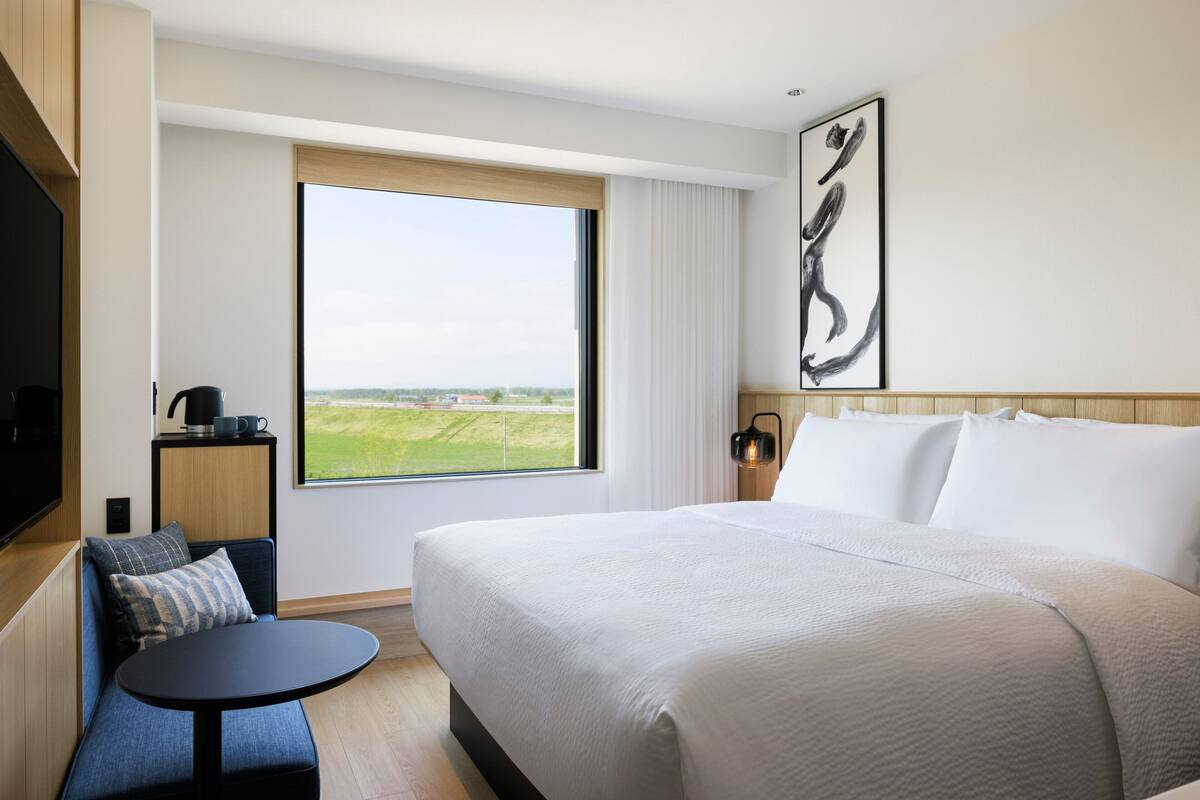 A hotel room with a large bed, a small round table, a sofa, a TV, a window with a view of the countryside, and a painting on the wall.