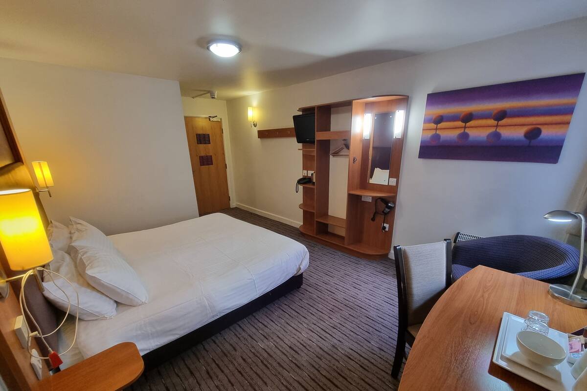 Une chambre d’hôtel avec un lit, une table de chevet, un bureau, une chaise et un tableau au mur.