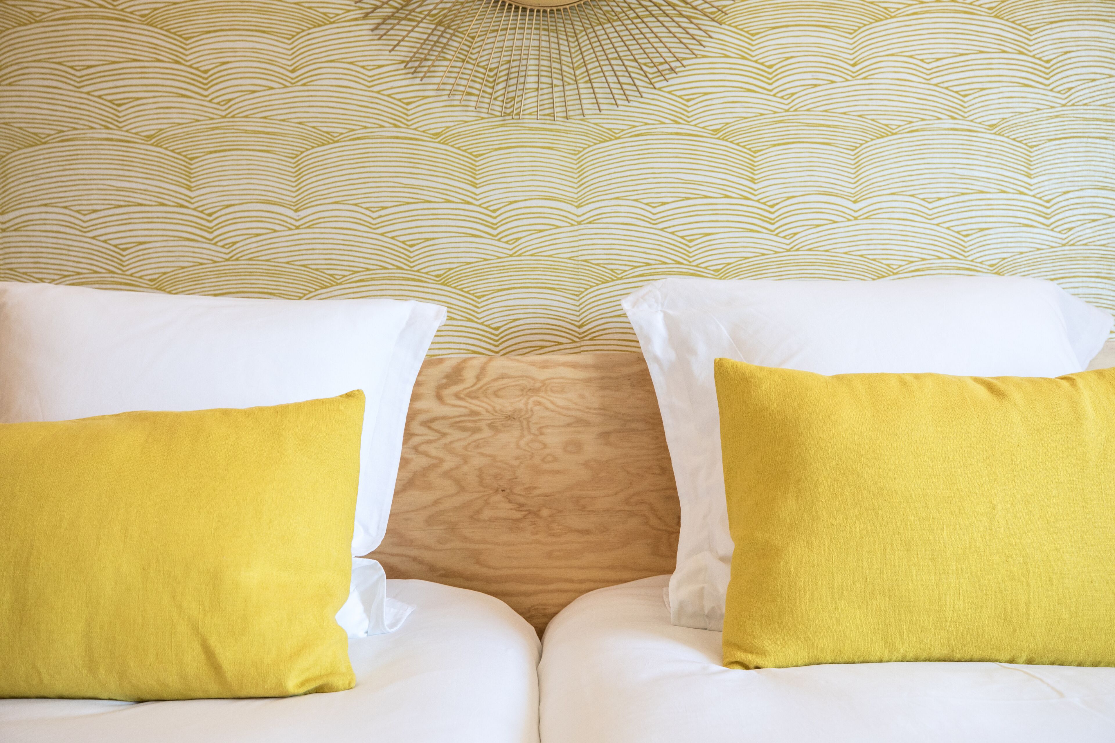 Una cama con almohadas blancas y amarillas, un papel tapiz estampado y un cabecero de madera.