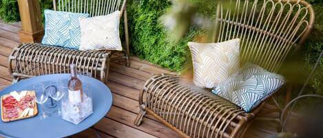 Wicker chairs with patterned cushions on a wooden deck, a table with a bottle and glasses, and a plate of food.