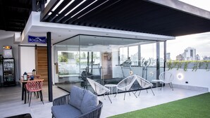 A modern outdoor seating area with a café sign, glass walls, and a view of the cityscape.