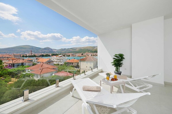 A balcony with a white table and chairs, a potted plant, and a view of a coastal town.