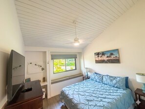 A bedroom with a bed, a desk, a chair, a ceiling fan, a window, and a painting on the wall.