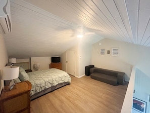 Een slaapkamer met een bed, nachtkastje, lamp, ventilator, bank en televisie.