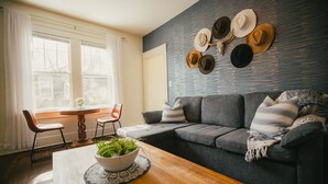 Een woonkamer met een grijze bank, een houten salontafel met een plant, een eettafel met stoelen, en een wand met hoeden.