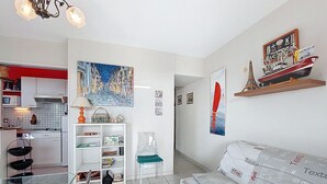 En moderne stue med sofa, sofabord i glas og bogreol. Der er et køkkenområde med hvide skabe og en mikrobølgeovn.