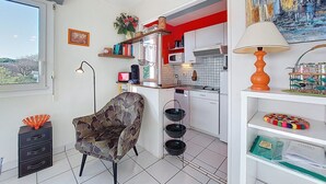 En kompakt opholdsstue med tekøkken, en stol, en lampe og en hylde med pynteting.