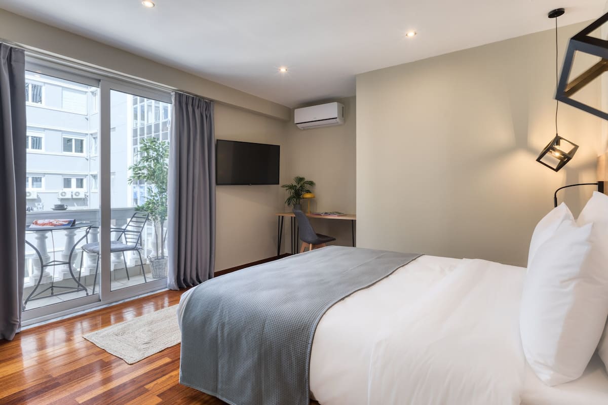 Habitación de hotel con cama, escritorio, televisión y balcón con vistas a edificios.