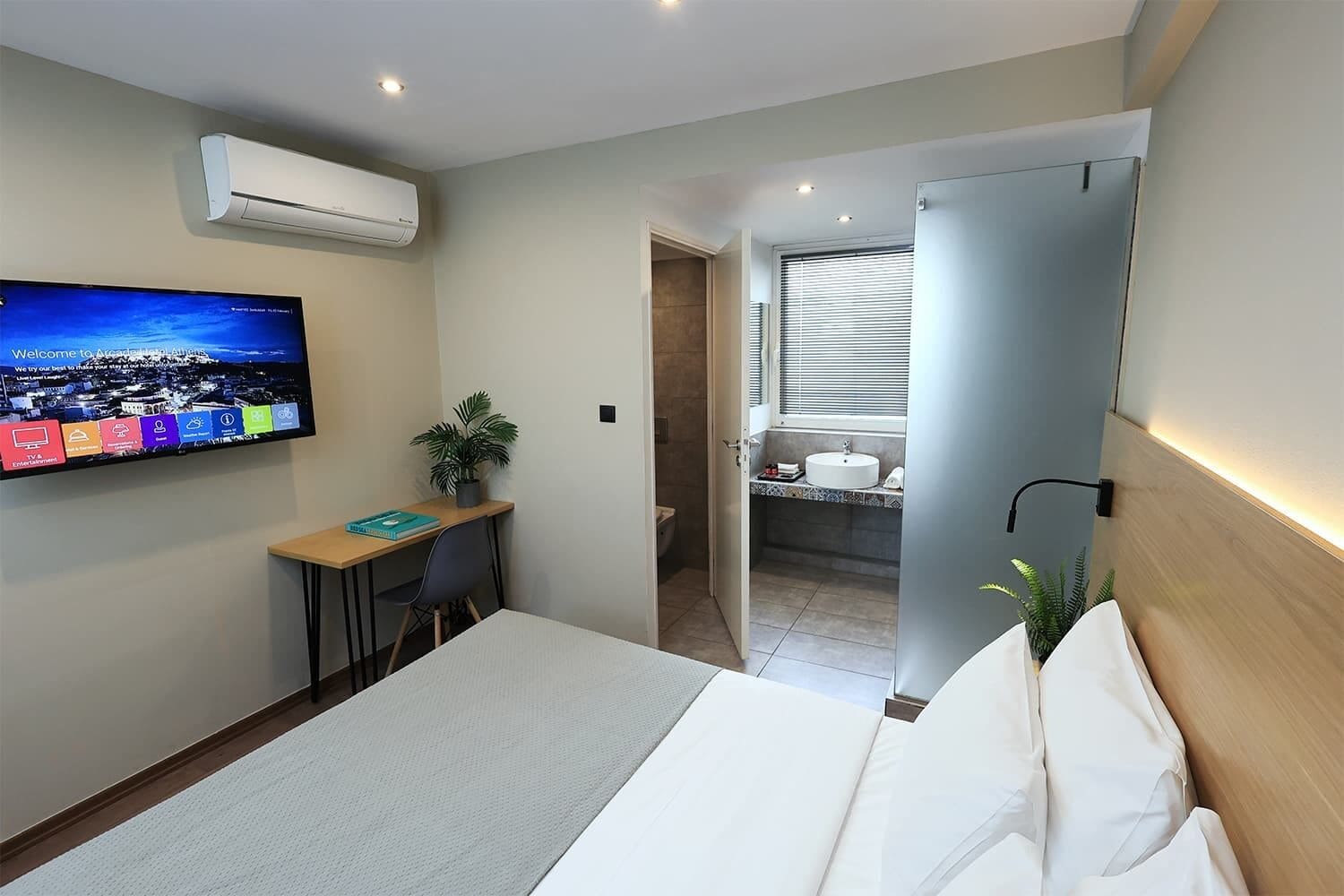 Een hotelkamer met een bed, een bureau met stoel, een aan de muur bevestigde televisie en een badkamer zichtbaar door een open deur.