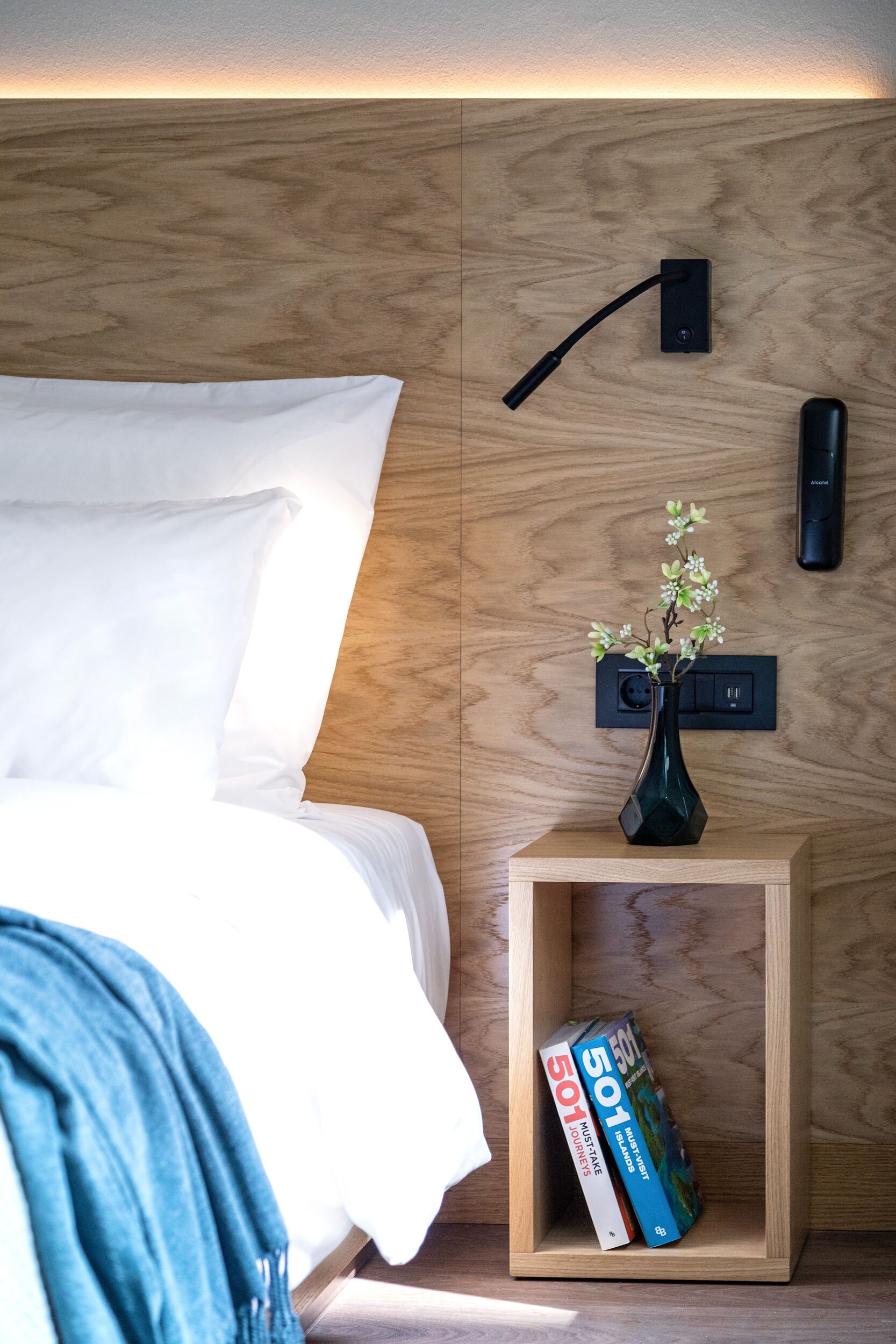 A neatly made bed with white linens, a bedside table with a vase of flowers, and two books titled '501' on a wooden nightstand.