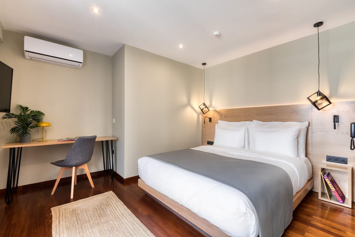 Un dormitorio moderno con una cama grande, un escritorio con silla, una planta pequeña y una mesita de noche con lámpara.