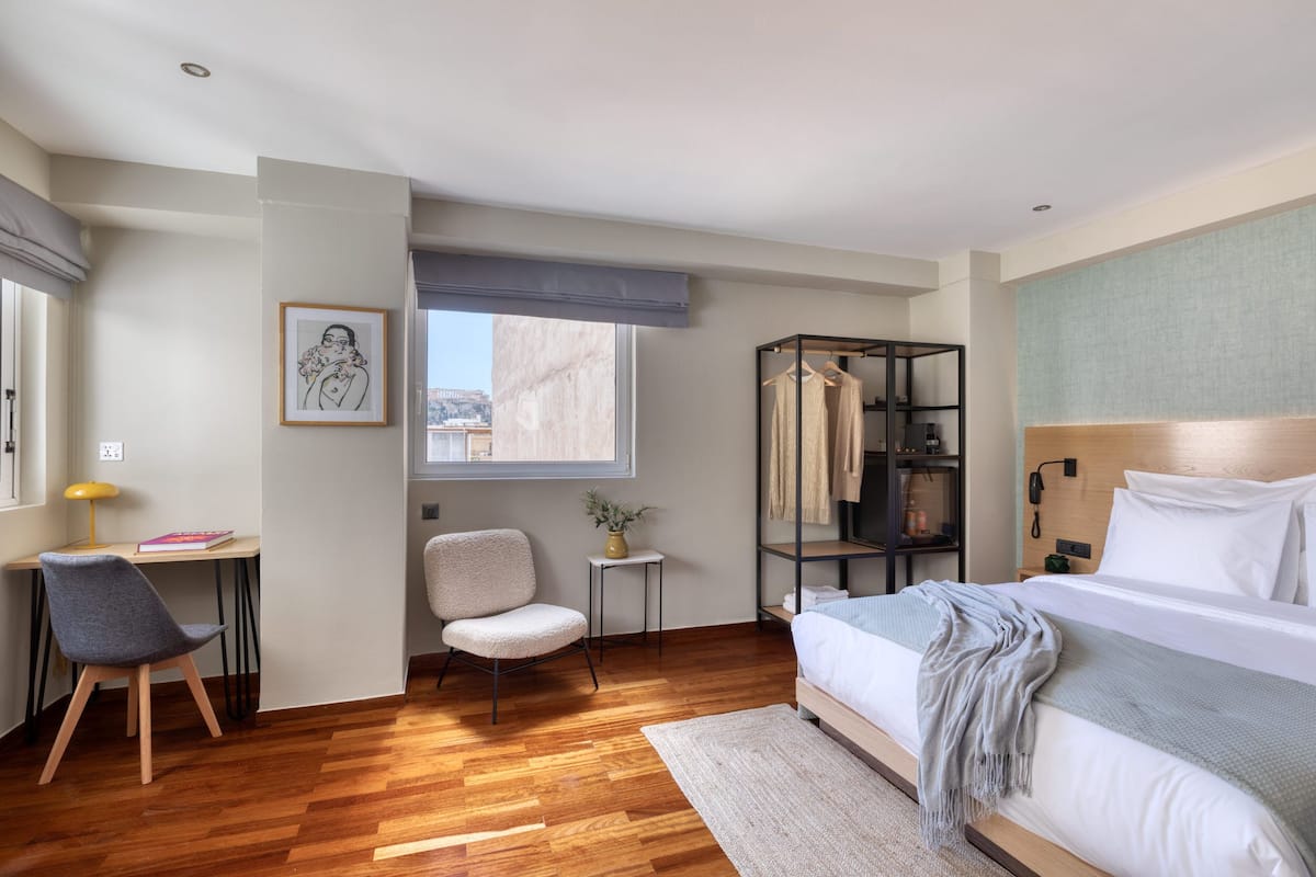 Una habitación de hotel moderna con piso de madera, una cama con ropa blanca, un escritorio con silla, una mesita con un florero y una ventana con vistas a edificios.
