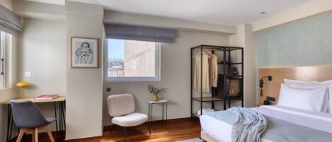 Una habitación de hotel moderna con piso de madera, una cama con ropa blanca, un escritorio con silla, una mesita con un florero y una ventana con vistas a edificios.