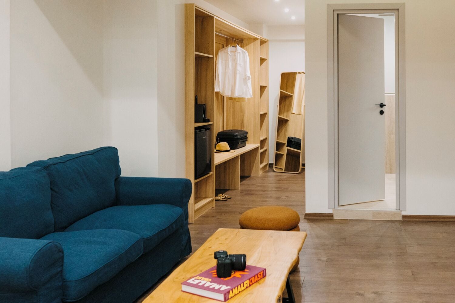 A modern living room with a blue sofa, a wooden coffee table with a camera and a book, and a hallway with wooden shelving and a door.