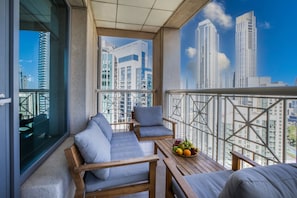 Ein Balkon mit einer Holzbank, Sesseln, einem kleinen Tisch mit Obst und Blick auf die Stadt.