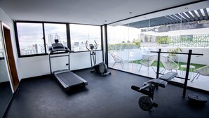 A modern gym with a treadmill, elliptical, and weight bench, large windows with a city view, and a balcony with outdoor seating.