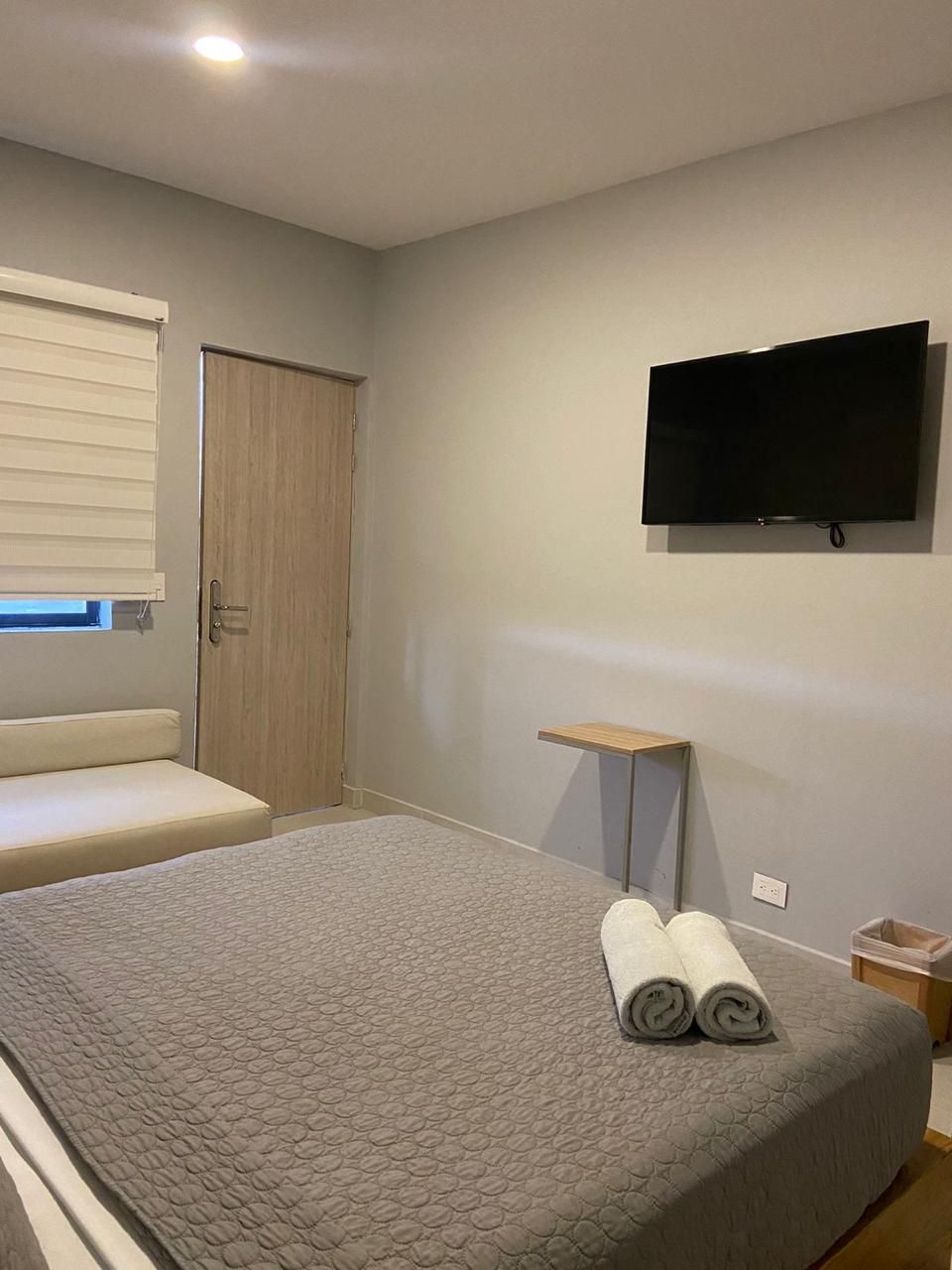 Un dormitorio con una cama, un televisor montado en la pared, una puerta de madera y un estante de madera pequeño.