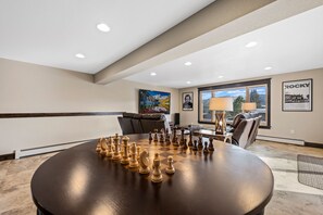Um quarto amplo com uma grande mesa redonda preparada para um jogo, uma área de estar aconchegante e vista através de uma janela grande.