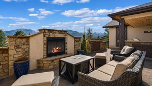 Patio esterno con pareti in pietra, un elemento decorativo a fuoco, mobili in vimini e vista sulle montagne.
