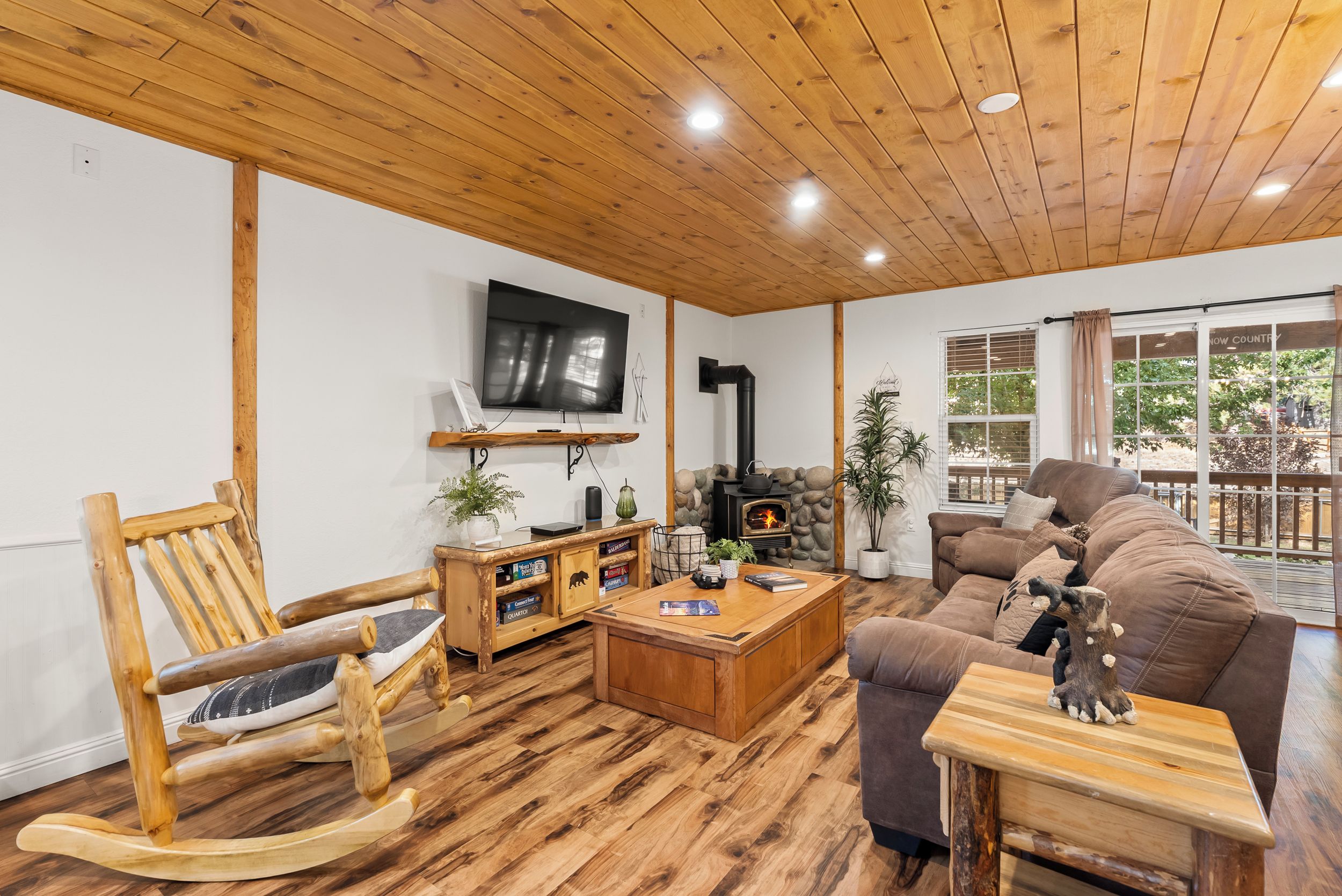 A cozy living room with a wooden ceiling, a flat-screen TV, a stone fireplace, and a large brown sofa.