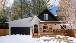 A cabin with a covered porch, a garage, and a snow-covered yard with picnic tables. - HOT TUB•MOVIE ROOM•GAME ROOM•DOGS ALLOWED•CENTRALLY LOCATED•FENCED IN PROPERTY (Big Bear Lake)