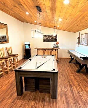 A pool table in a room with wooden flooring and ceiling, a bar area with a sign, and a billiards table.