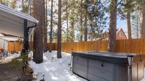 A backyard with a wooden fence, tall trees, and a hot tub. - HOT TUB•MOVIE ROOM•GAME ROOM•DOGS ALLOWED•CENTRALLY LOCATED•FENCED IN PROPERTY (Big Bear Lake)
