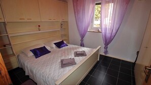 A bedroom with a bed, two pillows, a window with purple curtains, a wooden wardrobe, and a tiled floor. - Apartment for 3- 4 people in the center of Pula (Pula)