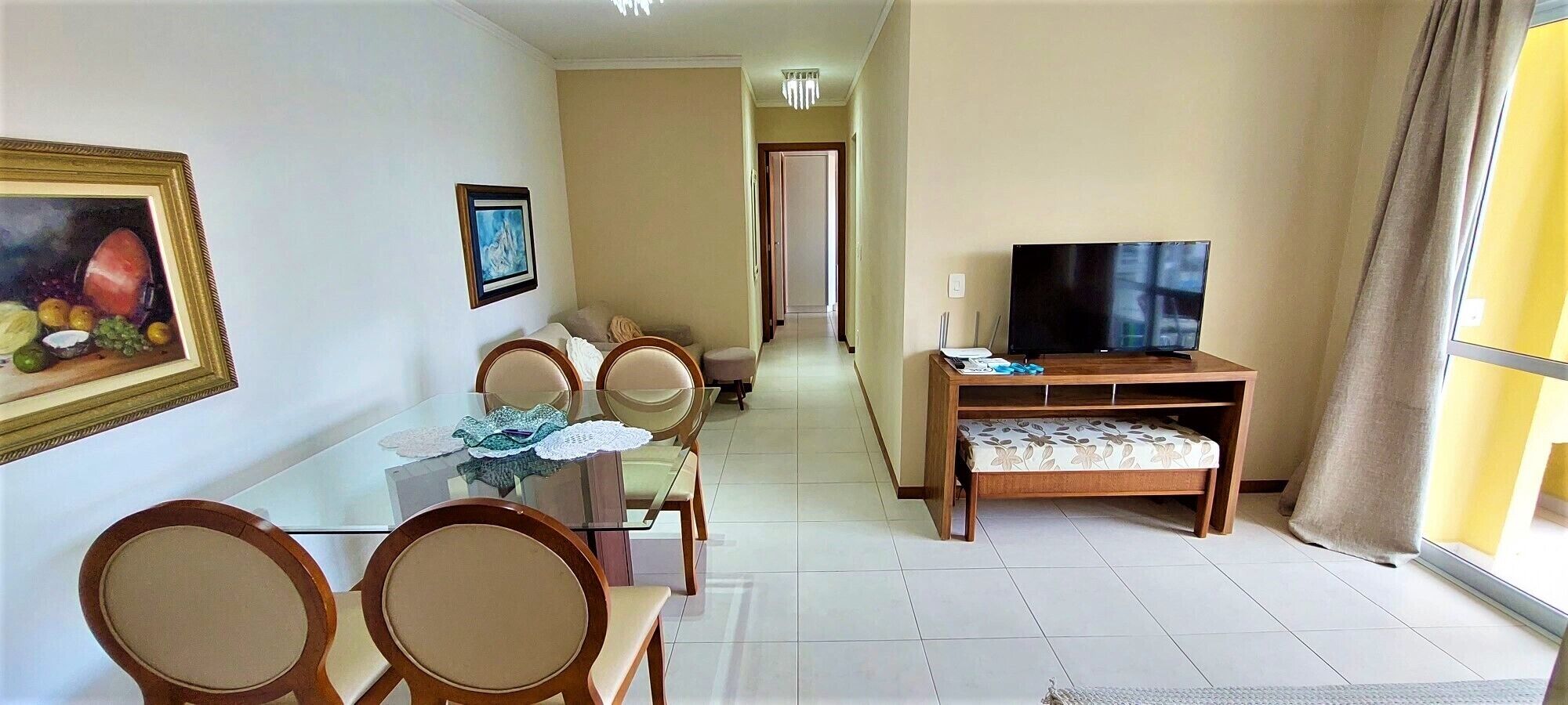 A dining area with a glass table and chairs, a television on a wooden stand, and a hallway leading to another room.