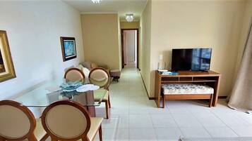 A dining area with a glass table and chairs, a television on a wooden stand, and a hallway leading to another room.