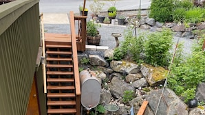 En træterrasse med en stenkø ved til en dør, omgivet af en have med forskellige planter og et klippearrangement.