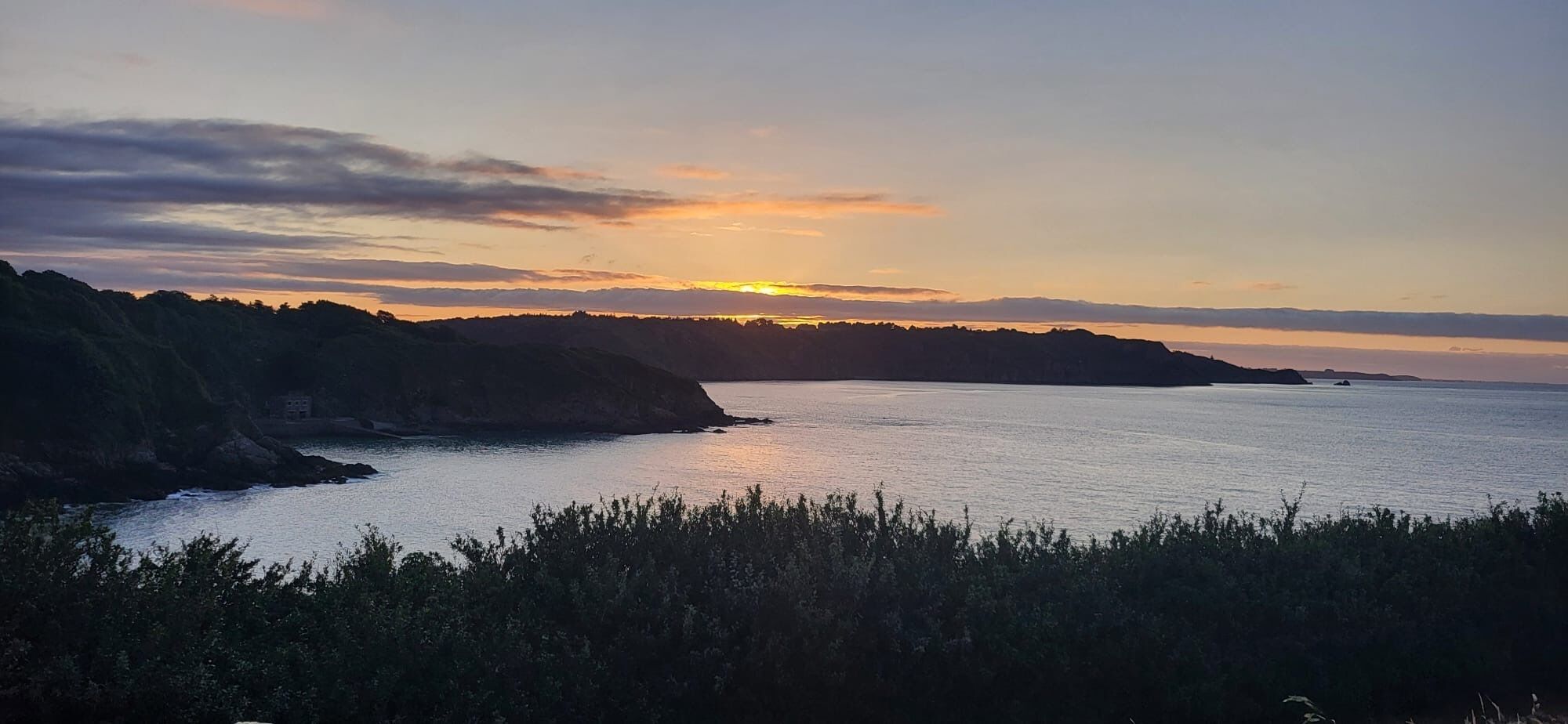 Un paysage côtier avec un coucher de soleil sur l’eau, des falaises et une végétation au premier plan.