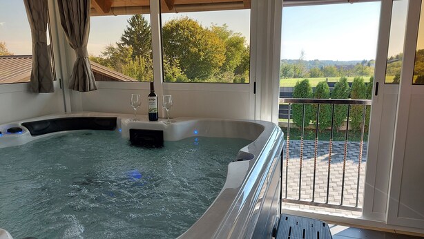 Uma banheira de hidromassagem com uma garrafa de vinho e copos, vista para árvores e um terraço de tijolos.
