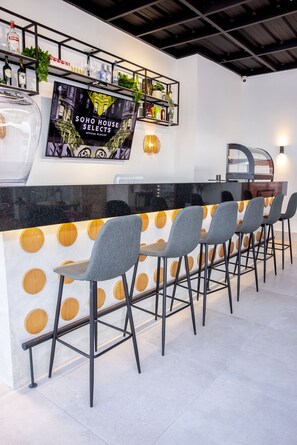 Un espace bar doté d’un comptoir noir, de quatre tabourets de bar gris et d’un écran de télévision diffusant une sélection de programmes intitulée « SOHO HOUSE SELECTS ».