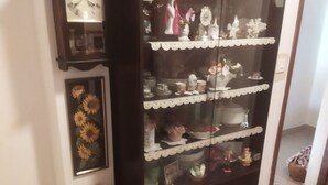 Een hal met een donker houten kastje met diverse decoratieve objecten. Links op de muur hangt een groot uurwerk en op de vloer staat een bloemstuk in een keramieken pot.