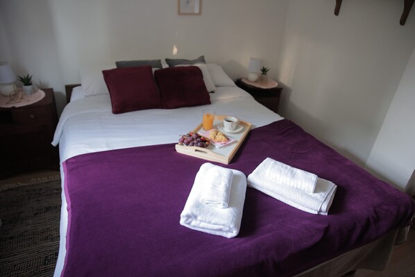 Una cama bien hecha con una colcha morada, almohadas blancas y dos toallas blancas dobladas. Hay una bandeja con una taza de café, un vaso de jugo de naranja y algunos pasteles sobre la cama.