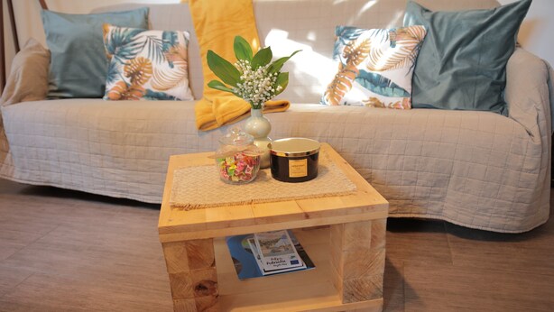 A wooden side table with a candle, a small vase with white flowers, and a decorative item on a beige sofa with patterned cushions. - Apartment Amigo - Two Bedroom Apartment with Terrace (Đurđevac)