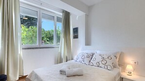 Ein Schlafzimmer mit Bett, Nachttisch, Fenster mit Vorhängen und Blick auf Grünfläche.