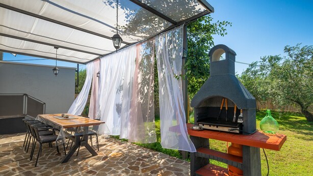 Eine Terrasse im Freien mit Steinboden, einem Holztisch mit Stühlen, einem Grill und einer Pergola mit leichten Vorhängen.